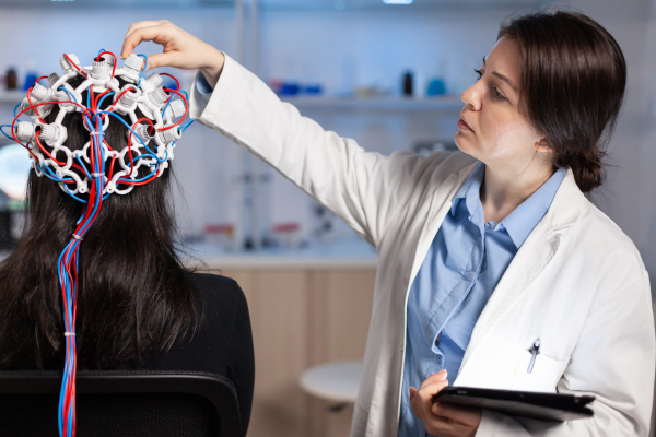 Back view of woman patient wearing performant eeg headset sitting on chair in neurological research laboratory while medical researcher adjusting it, examining nervous system typing on tablet.